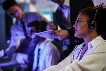 Cyber security analysts monitoring network threats in a blue-lit control room. IT specialists with headsets discuss digital safety and system defense data on a tablet device.