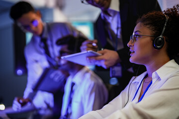 Cyber security analysts monitoring network threats in a blue-lit control room. IT specialists with headsets discuss digital safety and system defense data on a tablet device.