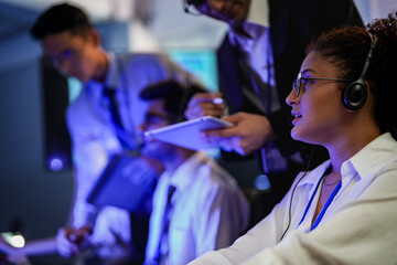 Cyber security analysts monitoring network threats in a blue-lit control room. IT specialists with headsets discuss digital safety and system defense data on a tablet device.