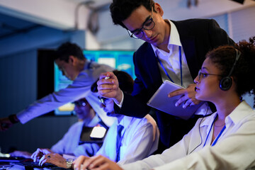 Cyber security analysts monitoring network threats in a blue-lit control room. IT specialists with headsets discuss digital safety and system defense data on a tablet device.