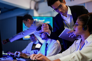 Cyber security analysts monitoring network threats in a blue-lit control room. IT specialists with headsets discuss digital safety and system defense data on a tablet device.