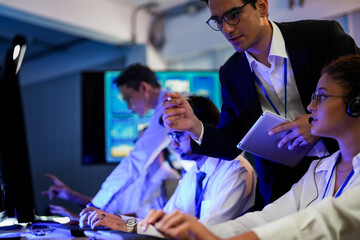 Cyber security analysts monitoring network threats in a blue-lit control room. IT specialists with headsets discuss digital safety and system defense data on a tablet device.
