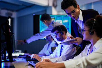 Cyber security analysts monitoring network threats in a blue-lit control room. IT specialists with headsets discuss digital safety and system defense data on a tablet device.