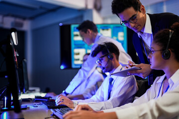 Cyber security analysts monitoring network threats in a blue-lit control room. IT specialists with headsets discuss digital safety and system defense data on a tablet device.