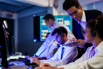 Cyber security analysts monitoring network threats in a blue-lit control room. IT specialists with headsets discuss digital safety and system defense data on a tablet device.