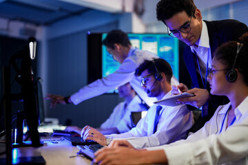 Cyber security analysts monitoring network threats in a blue-lit control room. IT specialists with headsets discuss digital safety and system defense data on a tablet device.