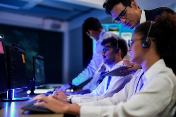 Cyber security analysts monitoring network threats in a blue-lit control room. IT specialists with headsets discuss digital safety and system defense data on a tablet device.