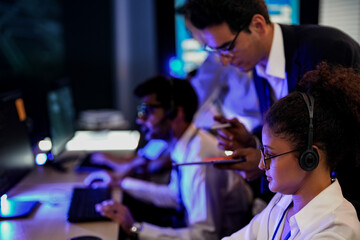 Cyber security analysts monitoring network threats in a blue-lit control room. IT specialists with headsets discuss digital safety and system defense data on a tablet device.