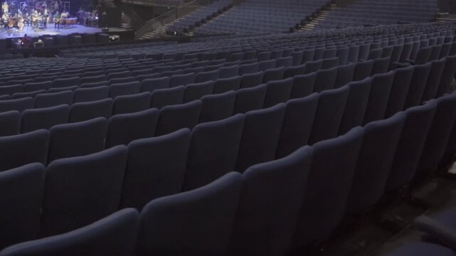 Auditorium filled with rows of empty seats near brightly lit stage with orchestra. Atmosphere of event rehearsal with focus on performers