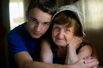 Young man gently hugging an elderly woman indoors, both looking at the camera, showing family connection, care, and quiet emotion in soft natural light.