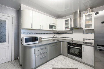 Fototapeta premium A modern L-shaped kitchen with white upper and grey lower cabinets, stainless steel appliances, built-in oven, microwave, curved countertop, frosted glass cabinets, tiled floor