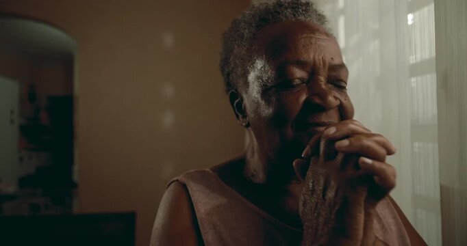 Elderly African American woman praying quietly by window, eyes closed, steadfast faith, comfort, spiritual grounding, and gentle resilience
