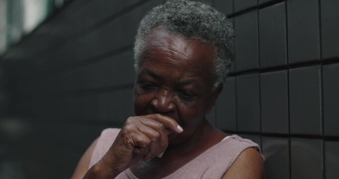 Elderly African American woman leaning against wall with hand near mouth, quiet preoccupation, worry, reflective pause, emotional depth, everyday dignity