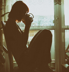 Silhouette einer nachdenklichen Frau sitzt am Fenster im Gegenlicht. Das Motiv vermittelt...