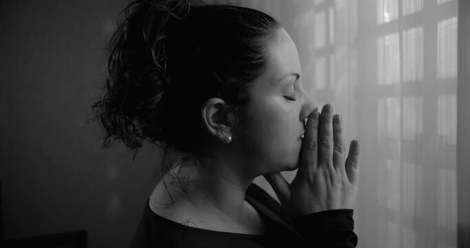 Woman standing by window in monochrome with hands pressed together in prayer, deep contemplation, spiritual focus, quiet surrender, intimate moment of faith