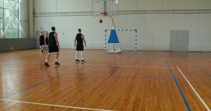 two basketball players come to practice and say hello to two teammates. a men's basketball team practices throwing the ball into the basket, the coach helps and suggests technique.