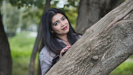 Bangladeshi girl peeking from behind tree trunk, looking at camera and smiling in park