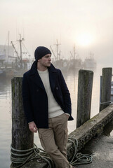 A thoughtful man in a peacoat and beanie stands on a pier in a misty harbor with fishing boats at sunrise. Maritime lifestyle and travel concept.