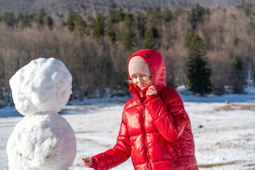 Young woman in a red winter jacket interacting with a snowman on a sunny winter day. Cozy seasonal...