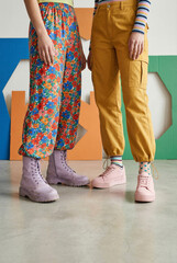Legs of two friends in colorful, trendy outfits. One in floral pants and lilac boots, the other in yellow cargo pants and pink sneakers. Youth fashion.
