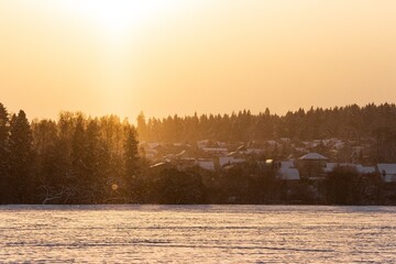 Naklejka premium Majestic golden light illuminates a charming winter village tucked amidst frosty trees and glistening snow. Delicate flakes dance, capturing the serene, crisp air