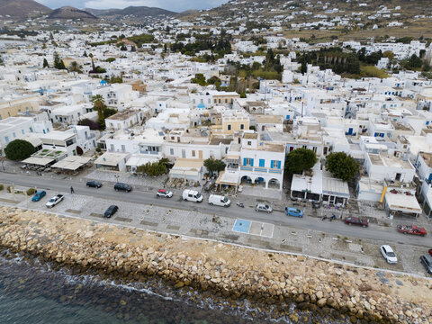 Aerial view of whitewashed buildings meet the azure sea along the coast, cars line the streets in a vibrant display of island life, Paros, Paros, Greece.