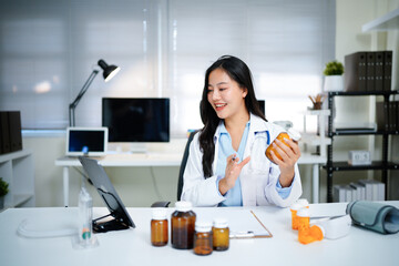 Professional asian female doctor in white gown sitting at desk with laptop, showing medicine bottle and explaining prescription during online consultation in hospital office