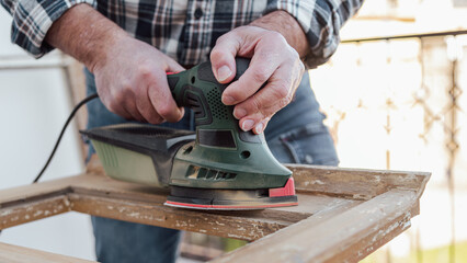 Craftsman. Adult carpenter using an electric sander to smooth an old wooden window. Construction industry, carpentry, housework do it yourself. Restoration.