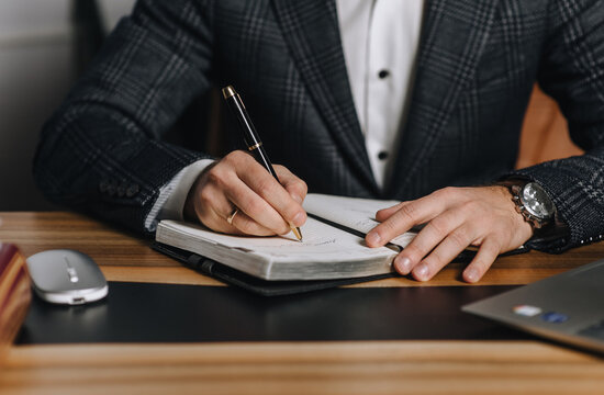 A close-up photo of an adult male businessman writing a business plan for a project in a notebook on a wooden office desk. Business concept, online documentation.