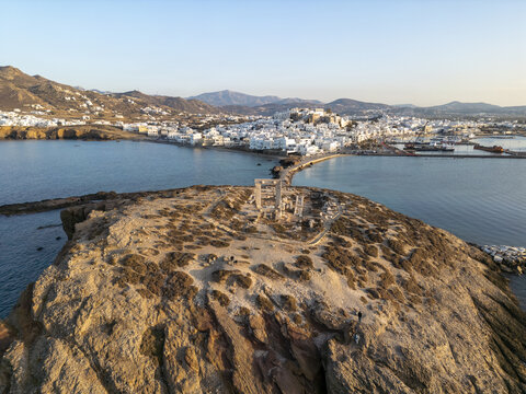 Aerial view of the ancient Temple of Apollo ruins standing proudly against the turquoise sea, connecting to the town's whitewashed buildings under a vast sky, Naxos, Cyclades, Greece.