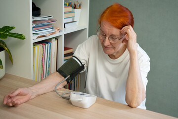 A 70-year-old elderly woman sitting at a table, attentively waiting for the result on the screen of her home blood pressure monitor. Concept of health monitoring and hypertension prevention.