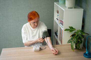 A mature woman is putting on the arm cuff of a digital blood pressure monitor at her home table. Getting ready for a health check. Focus on the action and device.
