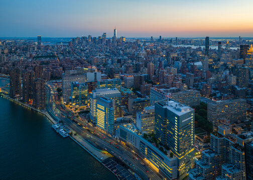 Aerial view of a sprawling cityscape where the East River reflects the fading light, skyscrapers pierce the horizon, and building lights twinkle like stars, New York, New York, United States.