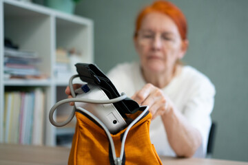 An elderly woman takes a blood pressure monitor from a small pouch or case. The device is the main focus, with the woman blurred in the background.