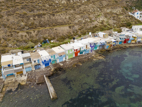 Aerial view of a vibrant row of colorful houses clinging to the rugged coastline, where the turquoise sea meets the quaint village of Klima, Milos, Greece.