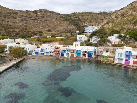 Aerial view of colorful boathouses stand in striking contrast against the turquoise waters and rugged hillside of Klima, Milos, Greece.