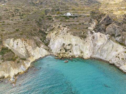 Aerial view of turquoise waters meet the rugged cliffs, where a solitary white building perches atop the hill, Mandrakia Beach, Milos, Greece.