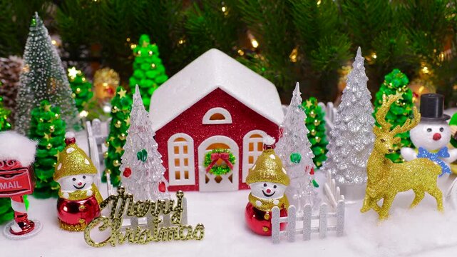 A Christmas village with a red house surrounded by snowmen and Christmas trees behind a fence and many Christmas trees and snowmen with a deer around against the backdrop of large Christmas tree branc
