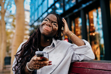 Thoughtful young Black woman with vitiligo holding smartphone, gazing up in contemplation, illustrating self-reflection, technology use, and digital lifestyle with authentic beauty