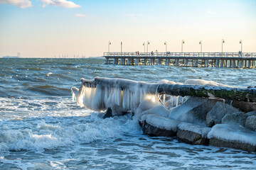 Winter pier at sunset with stormy sea, icy rocks and industrial skyline