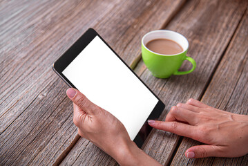 A woman's hands holding a digital tablet with a blank white screen next to a cup of coffee on a rustic wooden table.