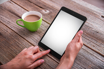 A woman's hands holding a digital tablet with a blank white screen next to a cup of coffee on a rustic wooden table.