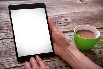 A woman's hands holding a digital tablet with a blank white screen next to a cup of coffee on a rustic wooden table.