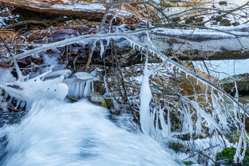Close-up of frozen icicles and ice formations on branches over a rushing stream. Detailed winter nature photography in a cold forest environment