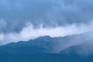 Clouds gather over mountain peaks in the early morning. The landscape shows a mix of gray and blue, with visible elevations. The scene is quiet and natural.