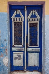 Wooden door haphazardly painted in indigo blue and white with neo-Classical ornate, on a mustard, pink and light blue faded wall. Tangier-Morocco-009
