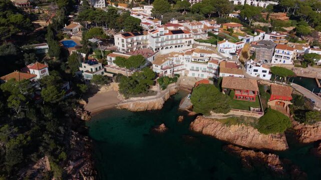 Drone footage ascending from the secluded cove of platja d'en malaret, revealing the picturesque coastal village of fornells with its traditional white houses nestled along the rocky coast
