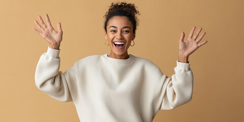 Excited woman with curly hair raising her hands and smiling broadly while wearing a cozy beige sweater on beige background.