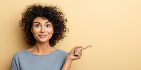 Surprised smiling woman with curly hair pointing to copy space against warm beige background.