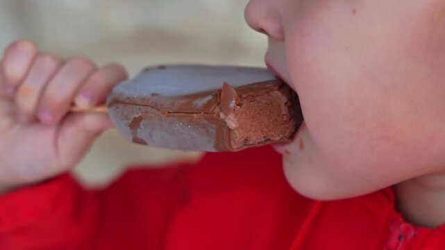 Tight closeup of a small child in a red jacket biting a chocolate ice cream bar on a wooden stick. Appetite and sweet treat concept, food and childhood, copy space